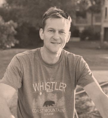 A man with short hair and sunglasses on his head smiles outdoors. He is wearing a t-shirt that reads Whistler Coast Mountains Canada. The photo has a sepia tone and a blurred background with greenery and buildings.