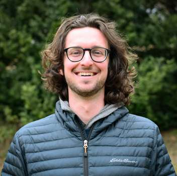 A man with wavy brown hair and glasses smiles while standing outdoors. He is wearing a dark puffer jacket and there are green bushes in the background.